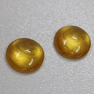 Op de afbeelding: Twee ronde, goudkleurige cabochons met een glanzende afwerking. De cabochons hebben een wervelend patroon van licht en donker goud, tegen een getextureerde, lichtgrijze achtergrond. Geschikt voor het maken van sieraden of knutselprojecten.
