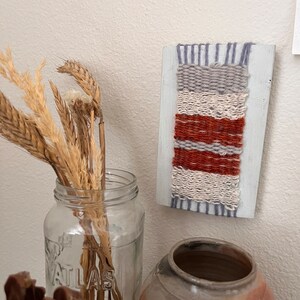 May include: A small woven wall hanging with a white background and stripes of gray, orange, and red yarn. The hanging is mounted on a white painted wooden frame.