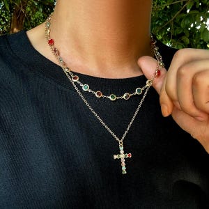 May include: Gold-toned necklace with two strands. The top strand features colorful gemstones, while the lower strand has a cross pendant adorned with matching gemstones. The necklace is worn against a black shirt.