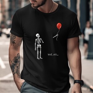 May include: A black t-shirt featuring a white graphic of a skeleton looking up at a red balloon floating away, with the text "Well, shit..." printed in white below the balloon. The design is centred on the chest of the short-sleeved garment.