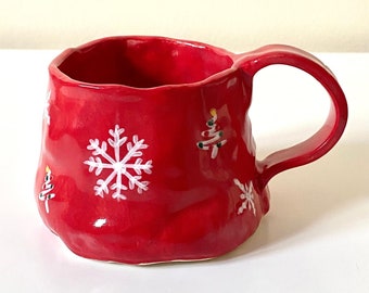 May include: A red ceramic mug with a textured surface. The mug features white snowflake and Christmas tree designs. It has a large handle and is suitable for hot beverages. The mug is a festive item for the holiday season.
