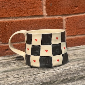 Puede incluir: Una taza de cerámica con base crema, con un diseño de tablero de ajedrez en blanco y negro. Pequeños corazones rojos están intercalados en el diseño. La taza tiene un asa curva y está colocada contra una pared de ladrillo y una superficie de madera.