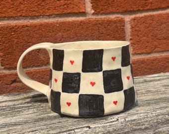 Puede incluir: Una taza de cerámica con base crema, con un diseño de tablero de ajedrez en blanco y negro. Pequeños corazones rojos están intercalados en el diseño. La taza tiene un asa curva y está colocada contra una pared de ladrillo y una superficie de madera.