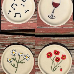 Puede incluir: Cuatro pequeños platos de cerámica blanquecina con diseños pintados a mano. Uno presenta notas musicales negras, otro una copa de vino tinto, un tercero margaritas blancas y el último amapolas rojas.