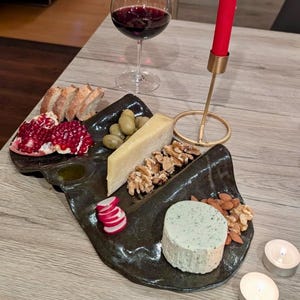 May include: A dark, irregularly shaped ceramic serving platter holds a selection of food including pomegranate seeds, green olives, sliced radish, a wedge of hard cheese, a round herb-flecked cheese, walnuts, almonds, and sliced bread.