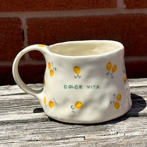 Könnte beinhalten: Cremefarbene Keramik-Tasse mit strukturierter Oberfläche, verziert mit gelben Zitronen-Illustrationen und grünen Blättern. Die Tasse trägt den grünen Schriftzug "DOLCE VITA" und hat einen gebogenen Henkel. Die Tasse steht auf einer verwitterten Holzoberfläche.