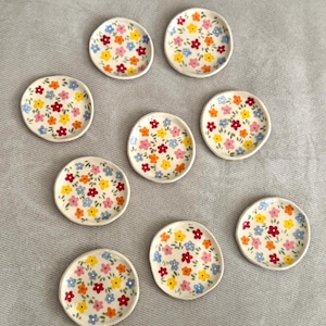 Puede incluir: Una colección de diez pequeños platos de cerámica blanquecina. Cada plato está adornado con un colorido estampado floral con flores rojas, amarillas, azules y naranjas. Los platos están dispuestos sobre una superficie gris claro.