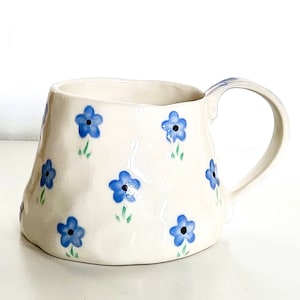 Könnte beinhalten: Weiße Keramiktasse mit einem blauen und grünen Blumenmuster. Die Tasse hat eine leicht unregelmäßige Form und einen Henkel.