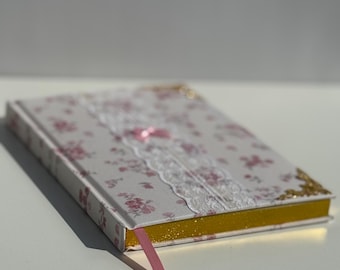 May include: A white journal with a pink floral pattern and lace trim. The journal has a gold edge and a pink ribbon bookmark.
