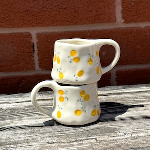 May include: Two white ceramic mugs stacked. Each mug features a handle and is decorated with yellow lemons and green leaves. The mugs are on a weathered wooden surface. The mugs are approximately 10 cm tall.
