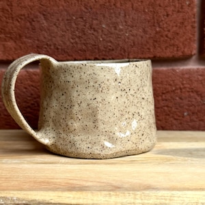 May include: A speckled beige ceramic mug with a rounded handle. The mug has a textured, slightly uneven surface.