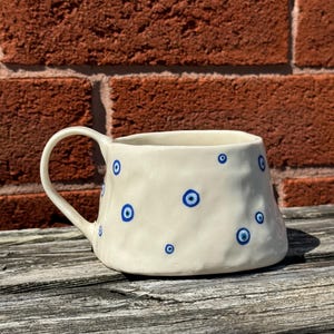 May include: White ceramic mug with a blue and white evil eye design. The mug has a handle and a slightly flared rim.