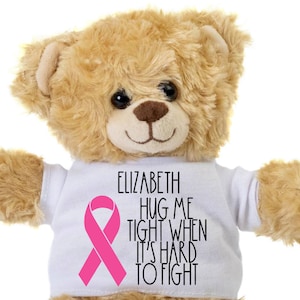May include: A beige teddy bear wearing a white t-shirt with a pink ribbon and the text "Elizabeth Hug Me Tight When It's Hard To Fight".