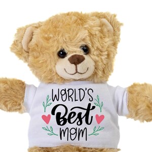 Könnte beinhalten: Ein hellbrauner Teddybär, der ein weißes T-Shirt mit dem Schriftzug "WORLD'S Best mom" in schwarzer Schrift trägt. Das Shirt hat grüne Blätterakzente und zwei rosa Herzen. Der Bär hat eine braune Nase und schwarze Augen.