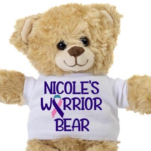 May include: A beige teddy bear wearing a white t-shirt with the text "Nicole's Warrior Bear" in purple lettering and a purple, pink, and teal ribbon.