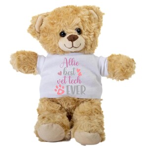 May include: A beige teddy bear wearing a white t-shirt with the text "Allie best vet tech EVER" in pink and gray lettering. The shirt also features a pink paw print with a white cross.