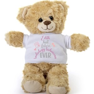 May include: A plush teddy bear with light brown fur wearing a white t-shirt. The shirt has the text "Edith best future radiology technician EVER" in pink and gray. The bear has a brown nose and black eyes.