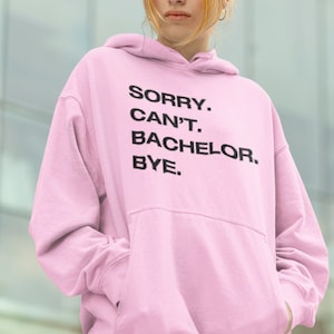 May include: Pink hoodie with the text "SORRY. CAN'T. BACHELOR. BYE." printed on the front.