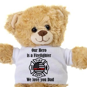 May include: A plush teddy bear with light brown fur wearing a white t-shirt. The shirt features a firefighter's emblem with the text "Our Hero is a Firefighter" and "We love you Dad". The bear has a friendly expression.
