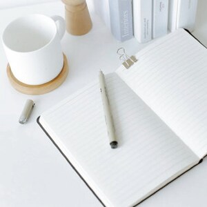 Puede incluir: Un cuaderno negro abierto con p&aacute;ginas rayadas, un bol&iacute;grafo plateado y una grapa plateada. El cuaderno est&aacute; sobre una superficie blanca.