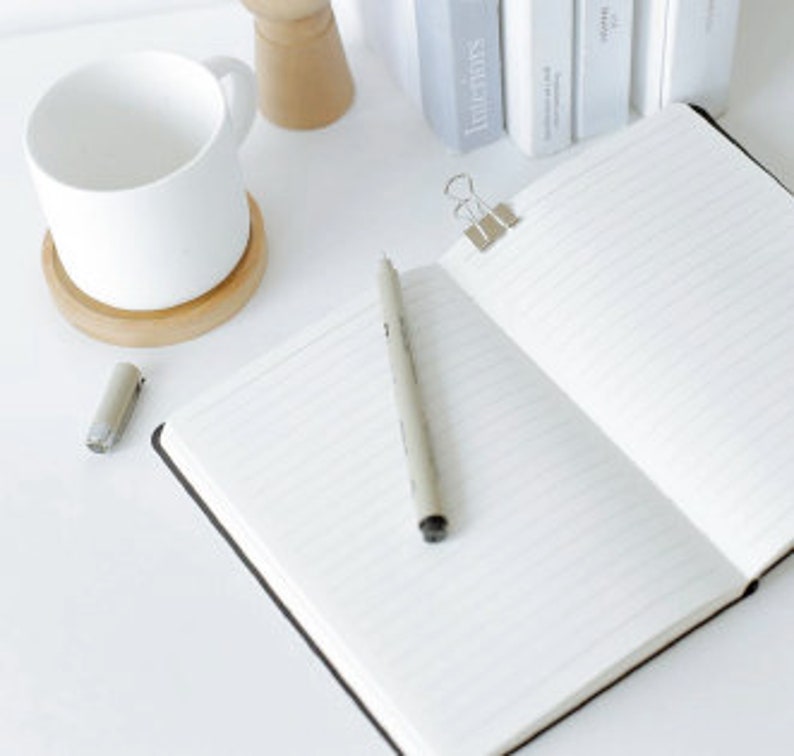 Puede incluir: Un cuaderno negro abierto con p&aacute;ginas rayadas, un bol&iacute;grafo plateado y una grapa plateada. El cuaderno est&aacute; sobre una superficie blanca.