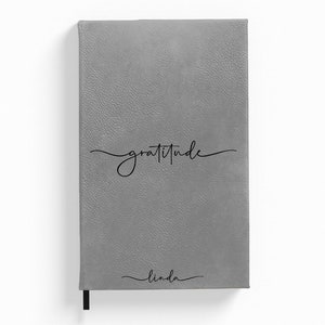 Puede incluir: Diario de imitaci&oacute;n de cuero gris con la palabra "gratitud" escrita en cursiva y el nombre "linda" escrito en cursiva en la parte inferior.