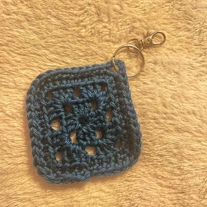 May include: A square, blue crocheted keychain with a gold-colored clasp and keyring. The keychain is made with a granny square pattern, featuring an openwork design in the center. The yarn appears to be a solid, dark blue color.