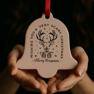 以下が含まれることがあります： 赤いリボンが付いた、薄いピンク色のベル型のクリスマスオーナメント。オーナメントには、鹿のイラストと「Wishing You a Very Scary Christmas - Merry Creepmas -」というテキストが黒で描かれています。