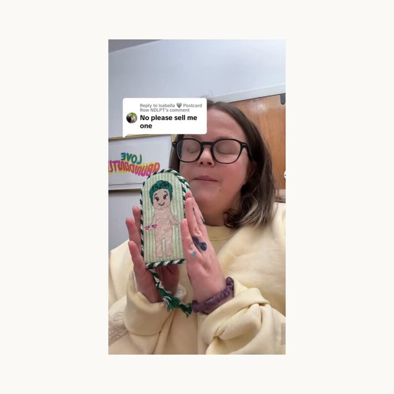 Może przedstawiać: Haftowany przedmiot w kształcie lalki z zielono-białym pasiastym tłem. Lalka ma zielone włosy i jest naga. Przedmiot jest obramowany zielono-białą obw&oacute;dką w paski i zielonym frędzlem. Widoczny jest tekst "No please sell me one".