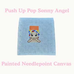 May include: A light blue needlepoint canvas featuring a Sonny Angel design. The design includes a cartoon character with a colorful hat and hands under its chin. The text "Push Up Pop Sonny Angel" and "Painted Needlepoint Canvas" are in pink.