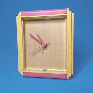 May include: A rectangular quartz clock featuring a pale yellow frame with pink horizontal accents at the top and bottom. The clock face has a light yellow grid pattern, pink numerals at the 12, 3, 6, and 9 positions, and pink clock hands.