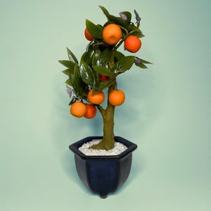 May include: A decorative artificial orange tree featuring bright orange spherical fruits and glossy dark green leaves. The tree has a textured brown trunk and is planted in a dark blue hexagonal ceramic pot filled with small white stones.