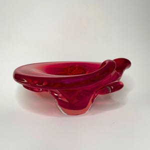 Peut inclure: Un bol en verre rouge rubis translucide et vibrant, présentant une forme organique asymétrique avec un bord épais et replié. La base est transparente et arrondie, soutenant le corps large et ondulant du récipient aux courbes fluides.
