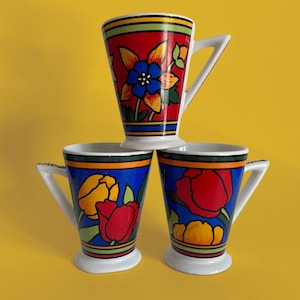 May include: Three ceramic mugs stacked in a pyramid formation. The top mug features a red background with a blue and yellow flower. The two bottom mugs have blue backgrounds with red and yellow tulip-like flowers. All mugs have white handles.