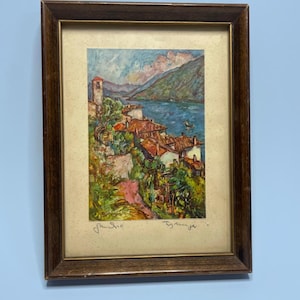 May include: A framed painting depicting a coastal village with terracotta-roofed houses, a tall bell tower, and lush greenery overlooking a blue body of water. The artwork is set within a dark brown wooden frame and features handwritten text.