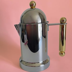 Peut inclure: Une cafetière en chrome poli dotée d'un corps cylindrique, d'un couvercle bombé avec un fleuron sphérique doré, et d'une poignée verticale ornée de capuchons sphériques dorés en haut et en bas. Le bec verseur est court et angulaire.