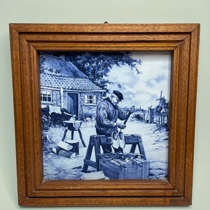 May include: A square wall art piece featuring a blue and white ceramic tile set in a medium-toned wooden frame. The tile depicts a person carving wooden clogs at a workbench in front of a cottage, with a windmill visible in the distant background.