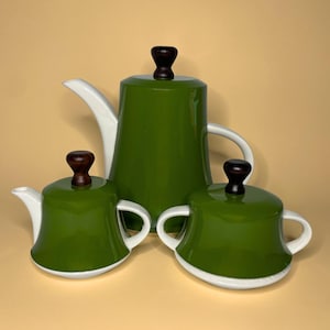 May include: A three-piece tea set featuring a tall coffee pot, a smaller creamer, and a sugar bowl. Each piece is finished in a glossy olive green glaze with white ceramic accents, including spouts and handles, topped with dark brown wooden knobs.