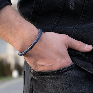 May include: A blue leather bracelet with a textured pattern and a silver clasp. The bracelet is worn on a wrist, with the person wearing a black denim jacket and black jeans. The bracelet is a stylish accessory.