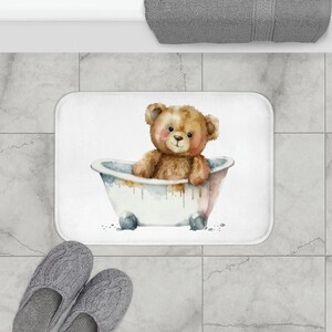May include: A white bath mat with a watercolor illustration of a brown teddy bear sitting in a white bathtub. The bear has a pink nose and brown eyes.