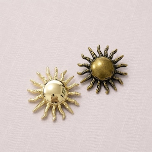May include: Two sun-shaped metal buttons or charms. One piece features a highly polished, reflective gold finish with a smooth center. The second piece has a matte, antique brass finish with a textured, darkened surface on the sun rays.