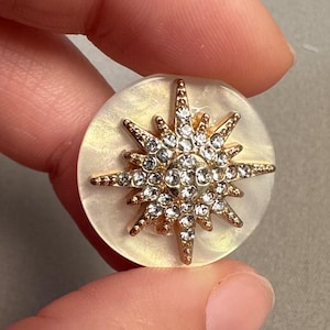 Puede incluir: Un botón redondo nacarado con un diseño de estrella dorada. La estrella está incrustada con brillantes pedrería transparente. El botón se sostiene entre dos dedos.