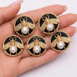 Peut inclure: Cinq boutons ronds dorés sur le thème de l'abeille, avec des fonds en émail noir et des accents de fausses perles. Les abeilles ont des ailes et des corps détaillés. Les boutons sont tenus dans une main.