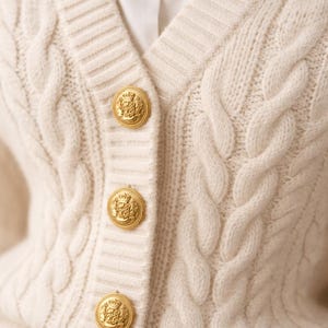 May include: Cream-colored cable knit cardigan with gold-colored buttons. The buttons are round with a detailed crest design. The cardigan has a V-neck and is made of a soft, textured material. A white shirt is visible underneath.