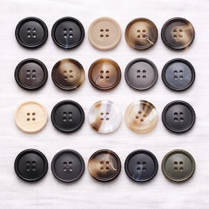 May include: A collection of twenty round buttons in diverse colours and patterns. The buttons feature solid black, beige, and marbled designs. Each button has four sewing holes and is arranged in a grid on white fabric.