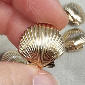 Könnte beinhalten: Nahaufnahme von mehreren goldfarbenen, muschelförmigen Verzierungen. Die Muscheln haben eine detaillierte, geriffelte Textur und ein glänzendes Finish. Das Bild zeigt die Muscheln in verschiedenen Positionen, eine davon wird zwischen Fingern gehalten.