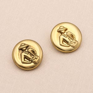 Op de afbeelding: Twee ronde goudkleurige metalen knopen met een reliëfontwerp van een figuur met een hoed met brede rand die een tas draagt. De knopen hebben een gladde, gepolijste afwerking en een licht gewelfde vorm met gedetailleerde afwerking.