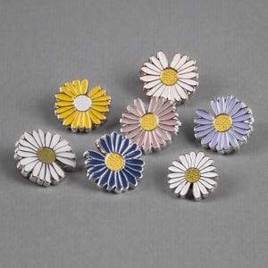 May include: A collection of daisy-shaped enamel pins in various colors, including white, yellow, blue, pink, and purple. Each pin has a silver-colored border and a yellow center, creating a vibrant floral design. The pins are arranged on a gray background.