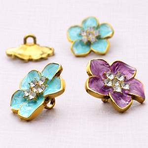 May include: Three flower-shaped buttons with gold-tone metal edges and centers adorned with clear crystals. Two buttons feature light blue enamel petals, while one button features purple enamel petals. Each button has a metal shank on the back.