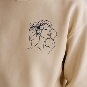 Puede incluir: Sudadera beige con un dibujo lineal negro bordado de un perfil de mujer con una flor en el pelo. El diseño está bordado en la parte superior izquierda del pecho de la sudadera. La sudadera tiene cuello redondo.